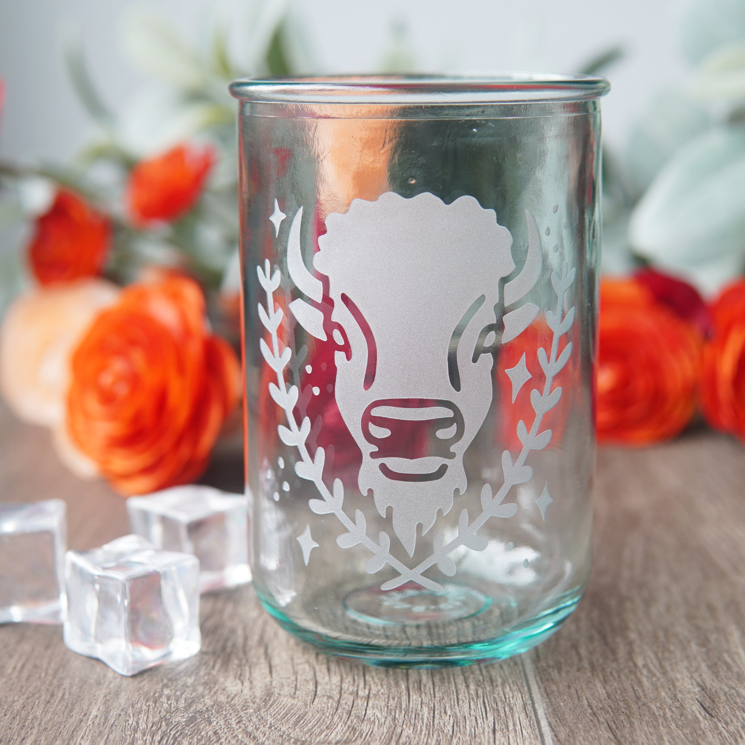 Buffalo engraved on a tall blue-tinged recycled glass cup, next to ice cubes and flowers