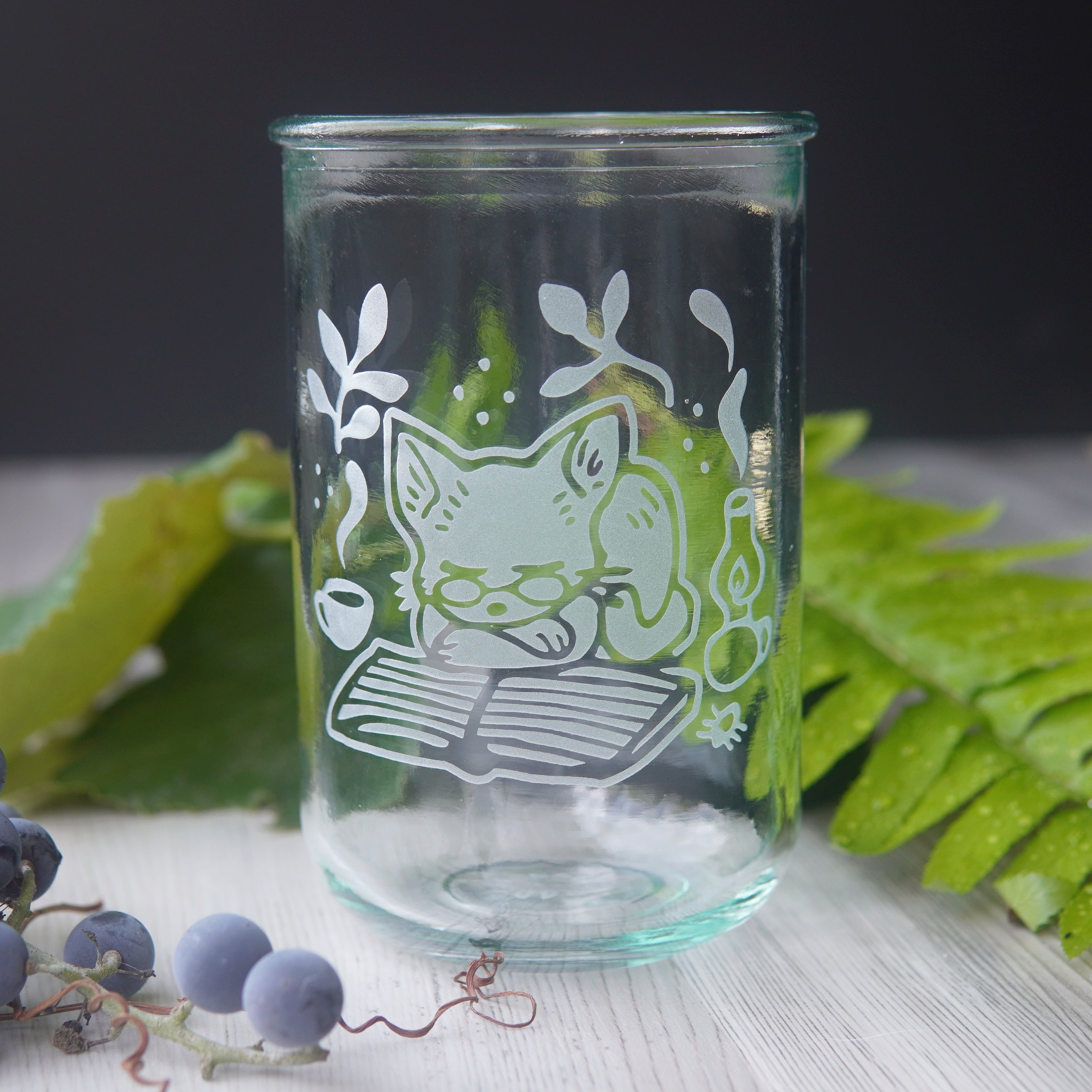 tall recycled glass cup engraved with a cat wearing glasses, sleeping on a book. There is a steaming mug and lit lantern next to the cat.