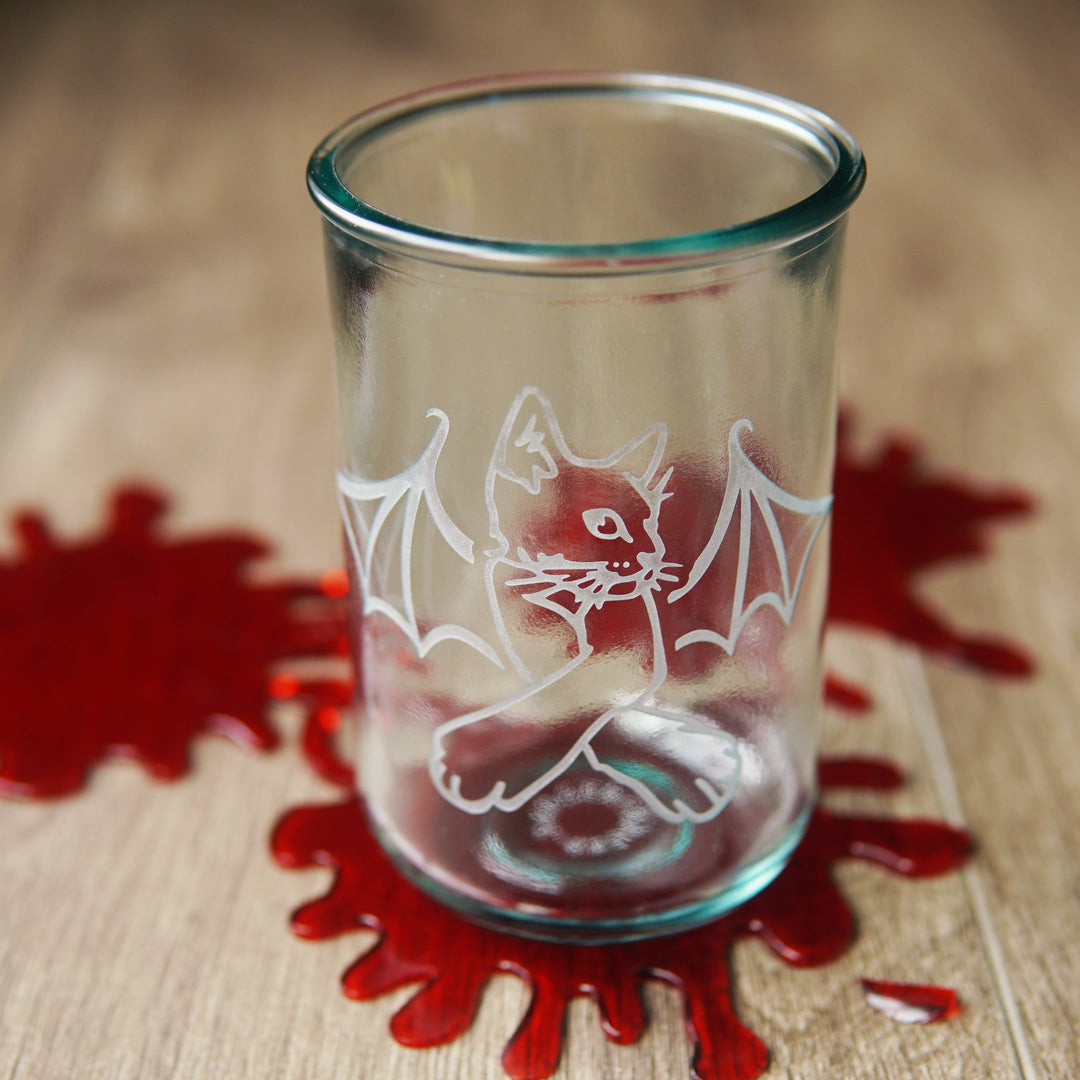 cat with bat wings engraved on a tall glass, with fake blood splatters on the table.