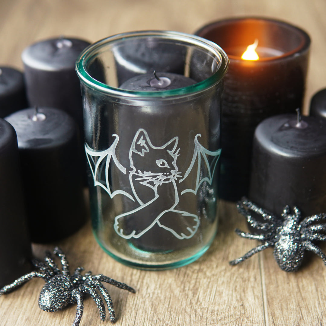 cat with bat wings engraved on a tall blue-tinged glass, with black candles all around, and glittery spiders