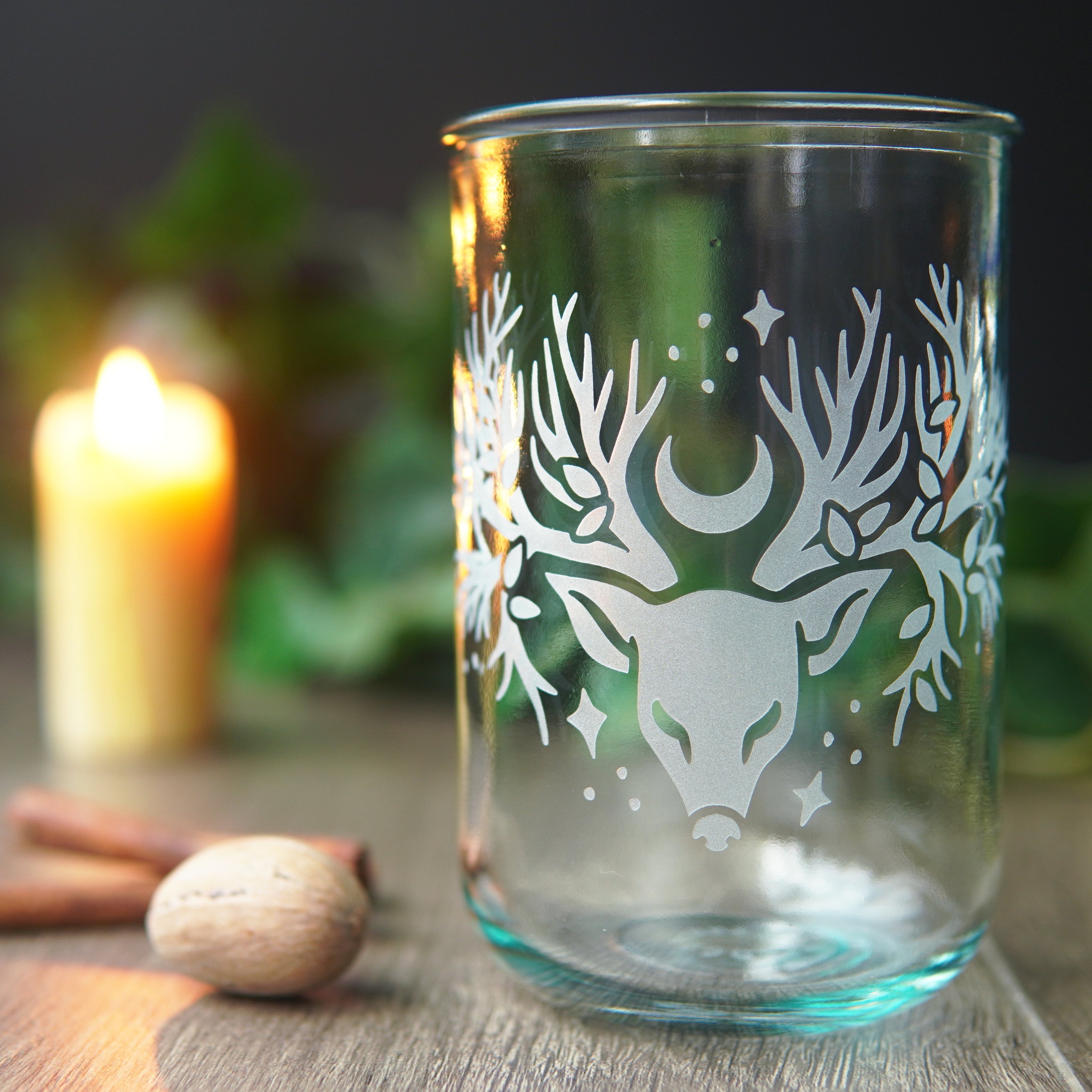 recycled glass cup engraved with a deer head with tree branches for antlers, sitting in front of a lit candle