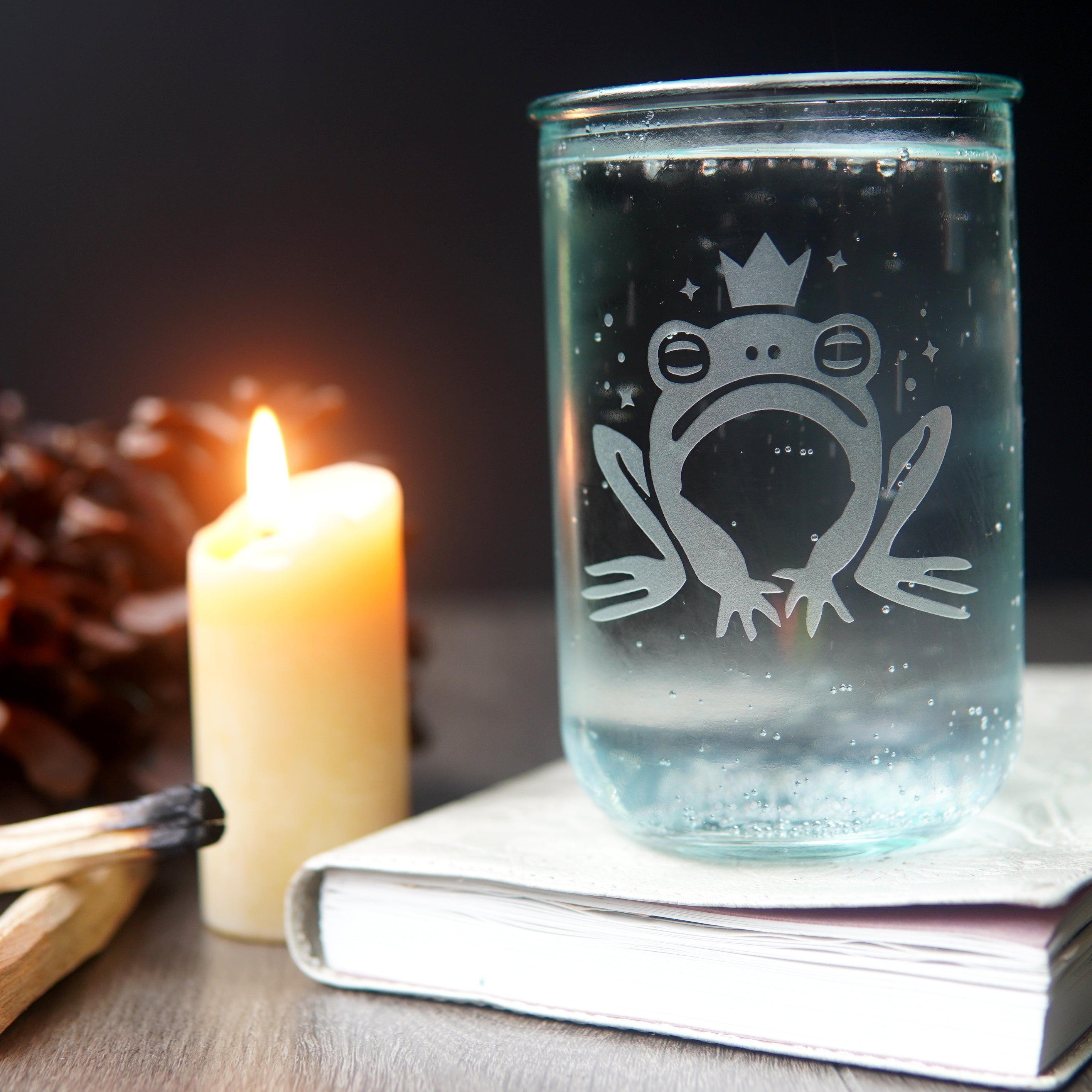 frog prince drinking glass full of seltzer, sitting on a book next to a lit candle