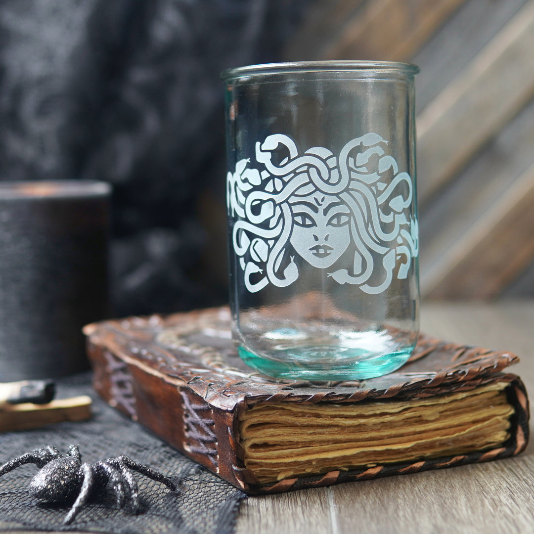 A clear glass tumbler with a white Medusa-style face design, displayed on top of an old book with a dark spider prop in a moody tabletop setting.