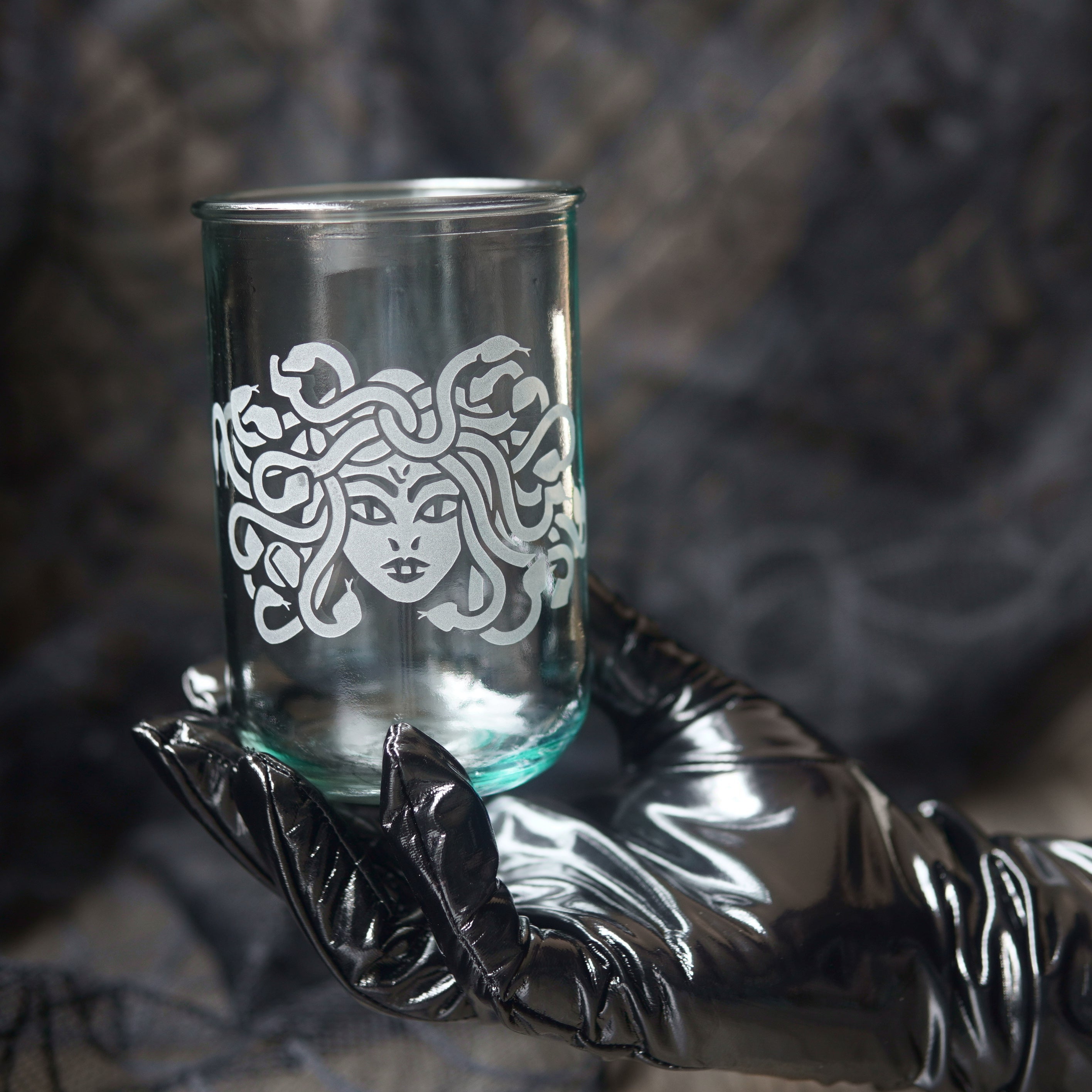 A clear glass tumbler with a white Medusa-style face and snakes motif, held in a shiny black glove against a dark textured background.