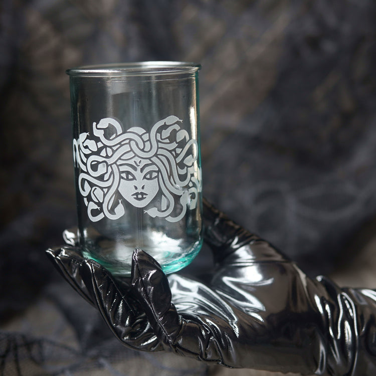 A clear glass tumbler with a white Medusa-style face and snakes motif, held in a shiny black glove against a dark textured background.
