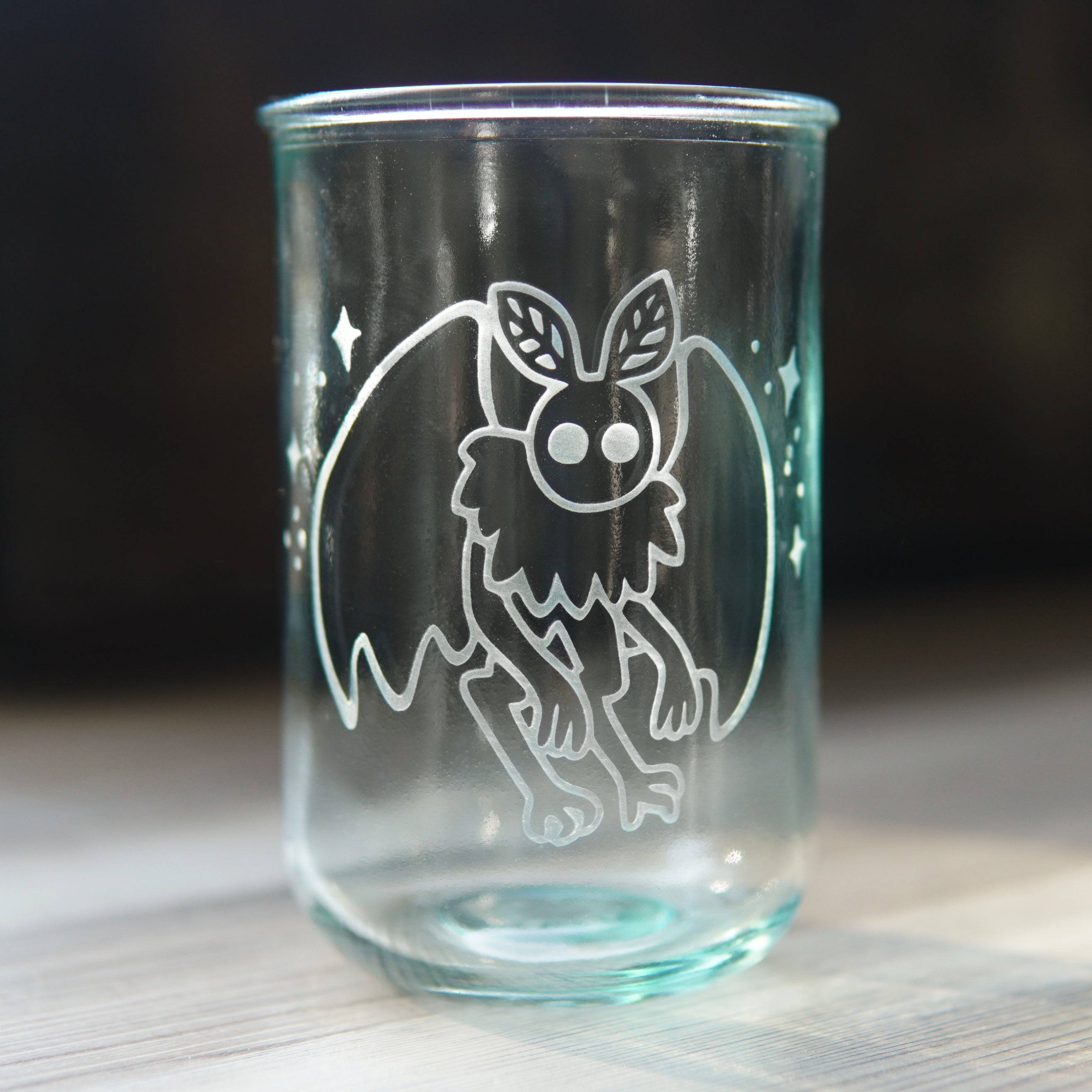 cute mothman surrounded by stars engraved onto a bluegreen-tinged tall glass