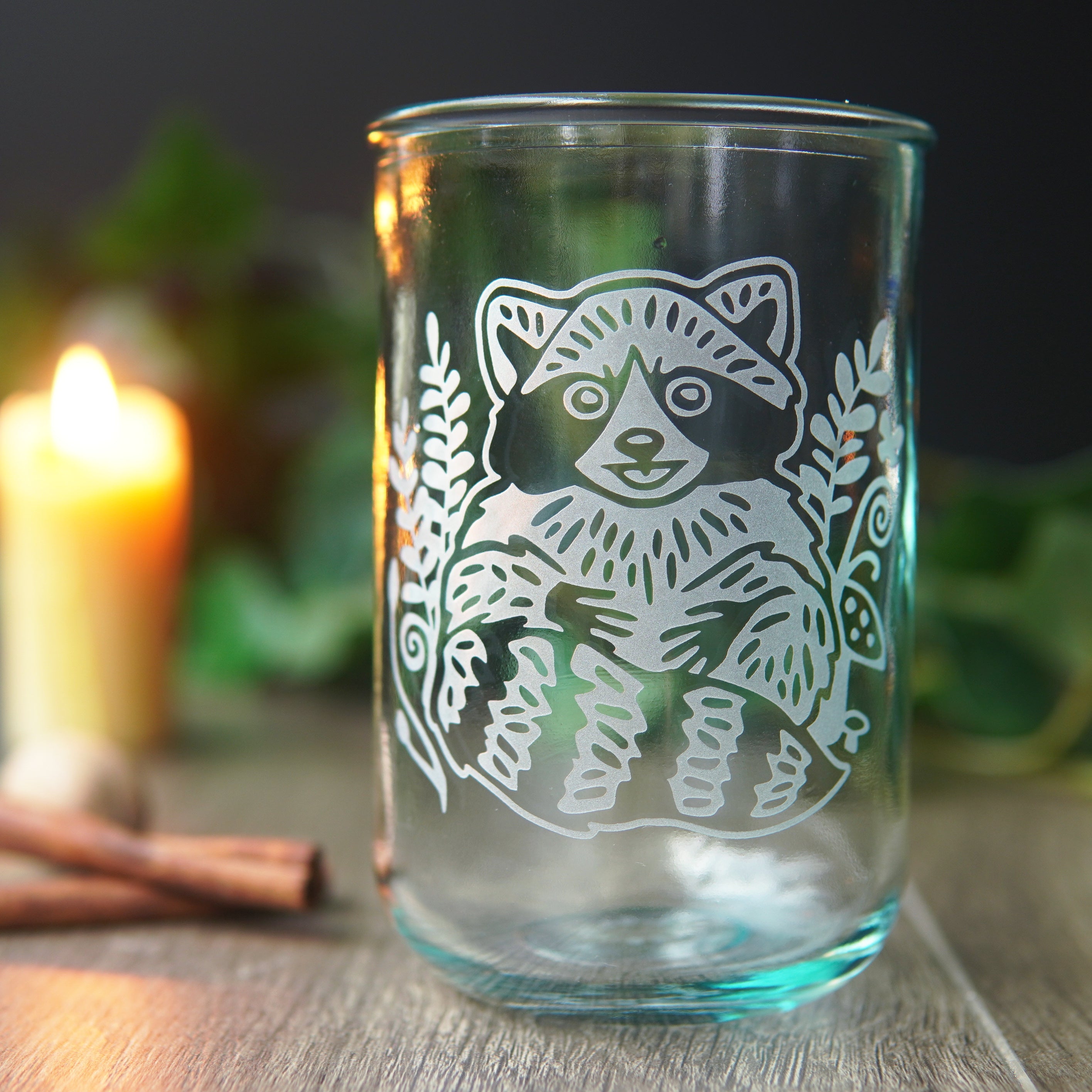 cute raccoon engraved on a bluegreen-tinged tall glass in front of a lit candle, cinnamon sticks, and greenery