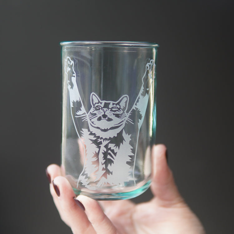 top half of a tabby cat with its arms stretched up and mouth slightly open engraved onto a tall glass, held in a hand