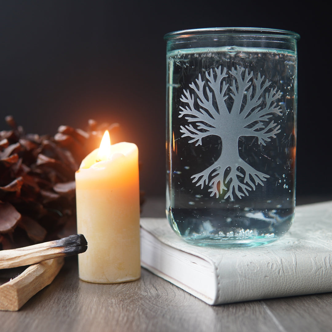 Tree etched on a tall glass full of seltzer, next to a lit beeswax candle, palo santo, pine cones, and a book