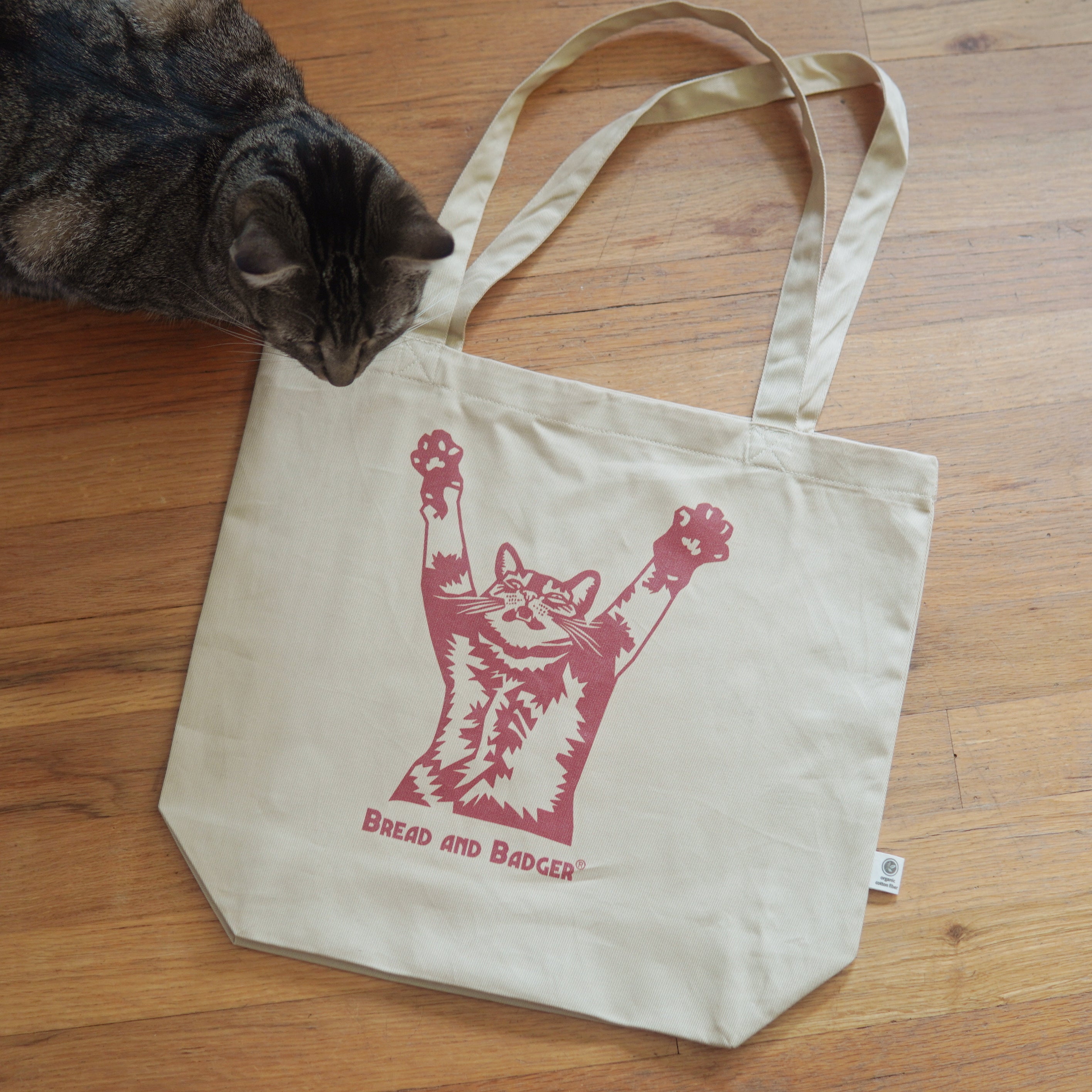 Beige tote bag with a red cat graphic and 'Bread and Badger' text on a wooden floor, with a cat sniffing the bag.