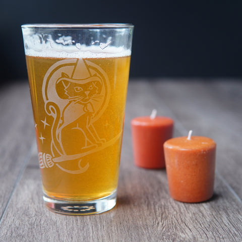 Witch Cat on a broom etched onto a pint glass next to fall votive candles and full of beer