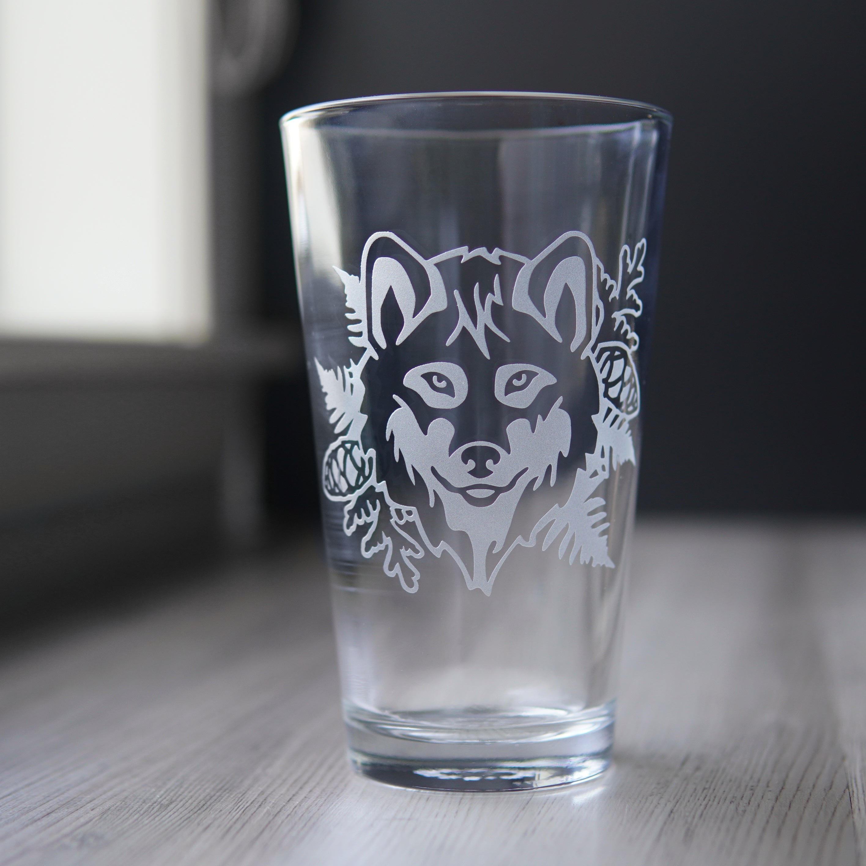 Pint glass engraved with a wolf's face, ferns, cedar branches, and pine cones. The wolf has a hint of a smile.